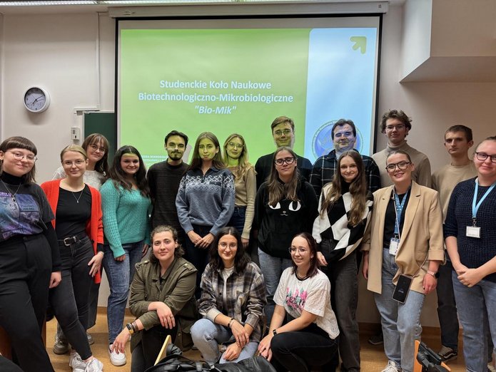Zdjęcie uczestników zebrania inaugarcyjnego SKN Biotechnologiczno-Mikrobiologicznego. Przedstawia 18 osób na tle slajdu zatytułowanego "Studenckie Koło Naukowe Biotechnologiczno-Mikrobiologiczne "Bio-Mik"".